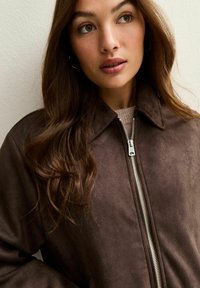 Chaqueta de ante sintético marrón con cierre de cremallera, cuello y textura suave. El fondo es neutral, lo que resalta el diseño de la chaqueta.