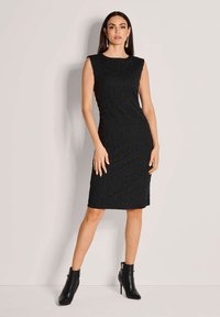 Schwarzes ärmelloses Kleid mit einem strukturierten Muster und einer figurbetonten Silhouette, kombiniert mit schwarzen Knöchelstiefeln, vor einem hellen Hintergrund stehend.