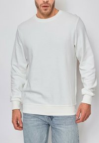Sweatshirt blanc à col ras du cou fabriqué en tissu texturé, avec des manches longues, des poignets et un ourlet côtelés ; associé à un jean bleu clair.