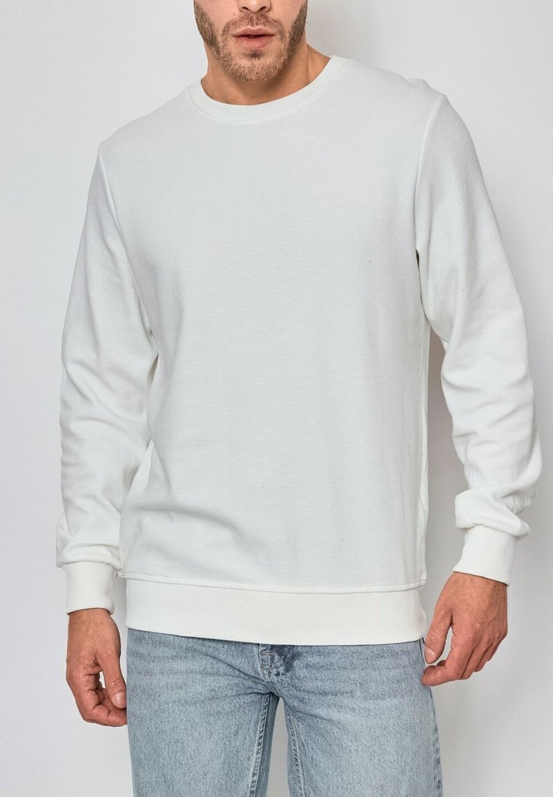 Sweatshirt blanc à col ras du cou fabriqué en tissu texturé, avec des manches longues, des poignets et un ourlet côtelés ; associé à un jean bleu clair.