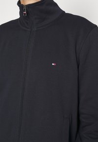 Primo piano di una persona che indossa una giacca nera con zip e un piccolo logo rettangolare rosso, bianco e blu sul petto.