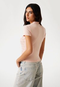 Růžový top s krátkým rukávem a přiléhavým střihem. Vlastní texturovaný vzor na levé straně a hladký zadní díl. Spárováno se světlými džínami.