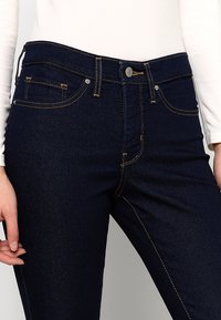 Mørkeblå denimjeans med gule stikninger, fem lommer og en metalknaplukning. Tætsiddende design med en glat tekstur.