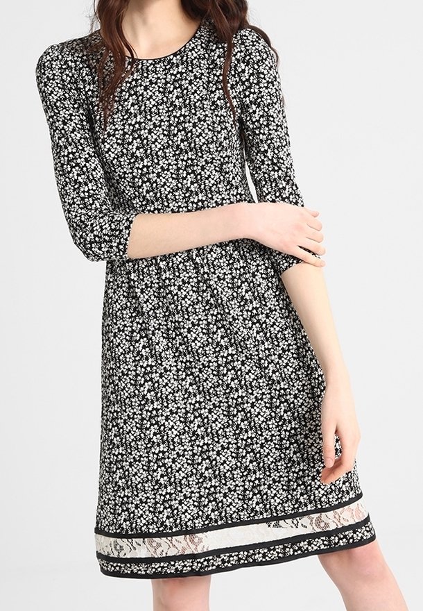 Femme portant une robe noire à petits motifs floraux blancs, manches trois-quarts et ourlet orné de dentelle, se tenant devant un fond uni.