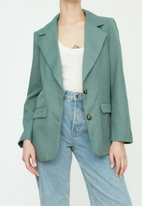Light green blazer with a structured collar, two buttons, and front pockets, paired with high-waisted light blue jeans. Smooth texture.