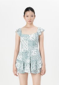 Jonge vrouw met haar opgestoken, gekleed in een mouwloze romper met een wit en groen bloemenpatroon, staande tegen een simpele witte achtergrond.