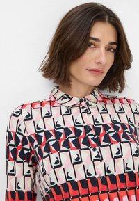 Camicia con bottoni caratterizzata da una stampa geometrica in rosso, blu navy e beige. Maniche corte, colletto e dettagli dei bottoni in evidenza. Tessuto morbido.