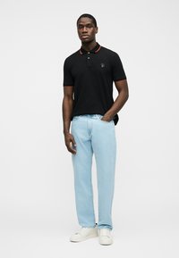 Άνδρας που στέκεται με το ένα χέρι στην τσέπη ανοιχτόχρωμου τζιν, φοράει μαύρο πόλο με κόκκινη λεπτομέρεια και λευκά αθλητικά παπούτσια, σε απλό φόντο.