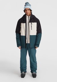 Mannen skikleding met een gelaagde jas in marineblauw, crème en teal, gecombineerd met bijpassende teal broek en zwarte skischoenen.