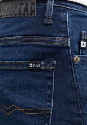 Tumši zili džinsa bikšu aizmugurējā kabata ar oranžām šuvēm, uz ādas uzlika un auduma etiķetes redzams "BIG STAR" zīmols.