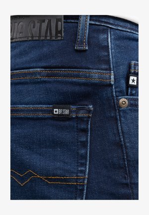 Tume sinised teksapüksid tagatasku oranži õmblusega, nahast plaastril ja kangasildil tähistatud "BIG STAR" logodega.