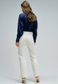 Blouse bleu marine à manches longues, associée à un pantalon blanc taille haute et des talons beiges. Tissu doux, coupe décontractée et détails minimalistes.