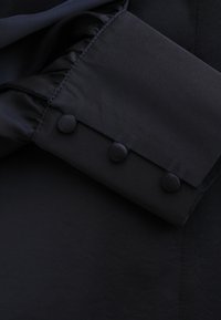 Czarna tkanina z marszczonym mankietem rękawa ozdobionym trzema guzikami obitymi tkaniną, podkreślająca gładki materiał i dopasowany krój.