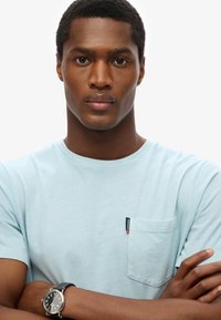 Camiseta de algodón azul claro con cuello redondo, mangas cortas y un bolsillo con una etiqueta. Se lleva un reloj de pulsera negro con una esfera de metal.