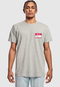 Camiseta gris de algodón, con cuello redondo y mangas cortas. Diseño notable de etiqueta roja con el texto "HELLO" en el lado izquierdo del pecho.