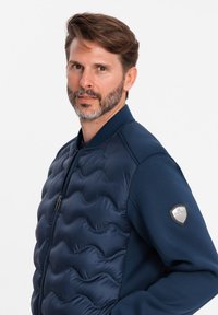 Veste bomber bleu marine avec un Devant matelassé présentant un motif de vagues, manches en tricot, et un badge en métal sur le bras gauche.