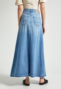 Light blue denim maxi skirt with a frayed hem, two back pockets, and a smooth waistband. Worn with black slide-on sandals.
