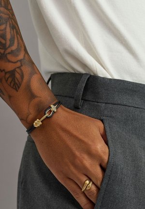 Sort flettet armbånd med guldfarvet hardware, inklusive en lås og dekorative detaljer. Båret på en hånd mod grå bukser.