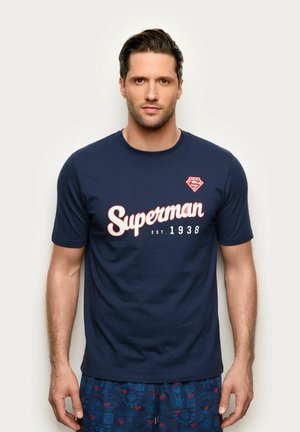 Mężczyzna zwrócony przodem, ubrany w granatową koszulkę Superman i dopasowane wzorzyste szorty na jednolitym jasnym tle.
