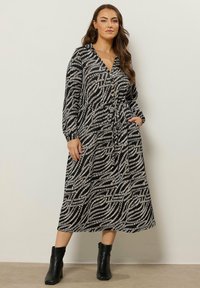 Kobieta stoi przy gładkiej ścianie, ubrana w midi sukienkę z długim rękawem w czarno-białe wzory z guzikami oraz czarne botki za kostkę.