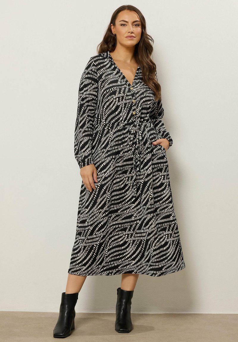 Kobieta stoi przy gładkiej ścianie, ubrana w midi sukienkę z długim rękawem w czarno-białe wzory z guzikami oraz czarne botki za kostkę.