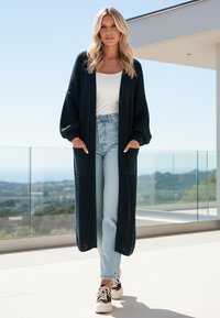 Długi granatowy kardigan z prążkowaną fakturą, otwarty z przodu, z kieszeniami po bokach, sparowany z jasnoniebieskimi, wysokim stanem dżinsami i wzorzystymi trampkami.