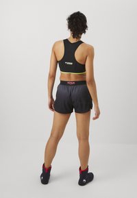 Schwarzer Sport-BH mit grünen Akzenten, kombiniert mit dunkelgrauen Shorts mit geometrischem Muster und rotem, markiertem Bund, abgerundet mit schwarzen Stiefeln.