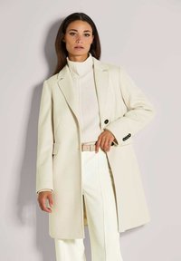 Beige wollen blend jas met een op maat gemaakt ontwerp, notch revers, twee klepzakken en zwarte knopen, gecombineerd met een roomkleurige coltrui en witte broek.