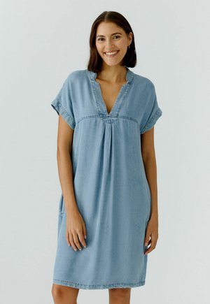 Lächelnde Frau mit schulterlangem dunklem Haar, die ein lockeres, hellblaues Jeanskleid mit kurzen Ärmeln und V-Ausschnitt trägt.