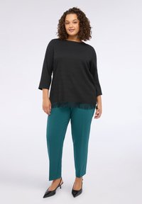 Schwarzes, strukturiertes Oberteil mit Fransen-Saum, kombiniert mit türkisfarbenen, schmal zulaufenden Hosen und schwarzen, spitzen Schuhen, die eine entspannte Passform und glattes Material präsentieren.