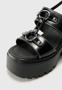 Black platform sandals with glossy patent finish, featuring wide straps, metallic accents, and contrast stitching on a textured sole.
