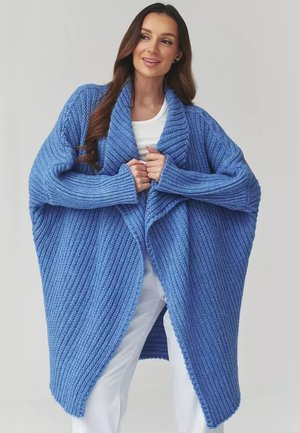 Kobieta w za dużym niebieskim dzierganym kardiganie na białym topie i spodniach, uśmiechająca się, z luźnymi, długimi, ciemnymi włosami na neutralnym tle.