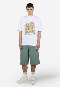 Weißes Baumwoll-T-Shirt mit einem farbenfrohen Obstmotiv und dem Schriftzug "LA DOLCE VITA", kombiniert mit Jeansshorts und weißen Turnschuhen.