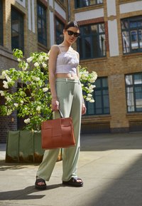Pantaloni larghi verde chiaro, top bianco corto, sandali neri e una borsa intrecciata marrone con manici neri. Ambientazione all'aperto con fiori.