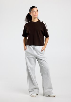 Vrouw staand met handen in haar zakken, gekleed in een zwart kortermig shirt met witte strepen en lichtgrijze wijde broekspijpen, met sneakers.