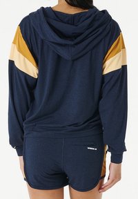 Un sweat à capuche marine à capuche et manches longues, orné de rayures dorées et crème sur les épaules, associé à un short marine avec une petite poche arrière.