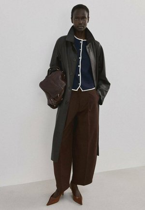 Lange persoon die een lange zwarte leren jas draagt, een marineblauwe, dichtgeknoopte cardigan, wijde bruine broek, bruine hakken, en een grote donkerbruine handtas vasthoudt.