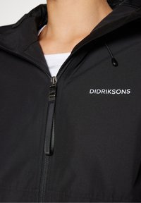 Gros plan d'une veste noire zippée avec le logo Didriksons sur la poitrine, portée par-dessus un haut blanc, avec une capuche et une languette de fermeture éclair.