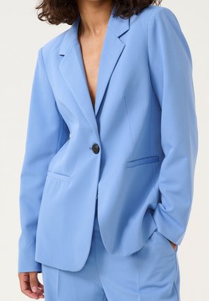 Femme portant un blazer bleu clair ajusté avec un bouton fermé et un pantalon assorti, main dans la poche sur un fond neutre.