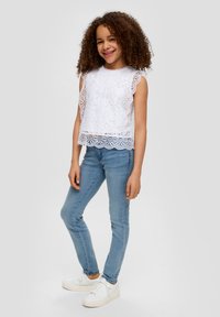 Biały top z koronką o falbaniastych rękawach, z teksturowanym kwiecistym wzorem, zestawiony z jasnoniebieskimi dopasowanymi dżinsami i białymi trampkami na platformie.