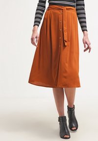 Rustfarvet midi-nederdel lavet af letvægtsstof, med frontknaplukning, bindebånd i taljen og et svajende design. Sorte ankelstøvler vist.