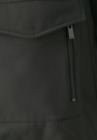 Svart tygjacka med en framficka med dragkedja, slät yta och en vikbar klaff vid fickan. Minimalistisk design med synliga sömmar.