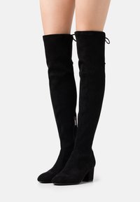Botas de ante negras sobre la rodilla con tacón de bloque. Presentan un cierre de cremallera en el lateral y punta redondeada. Cuentan con cordones en la parte superior para un ajuste seguro.