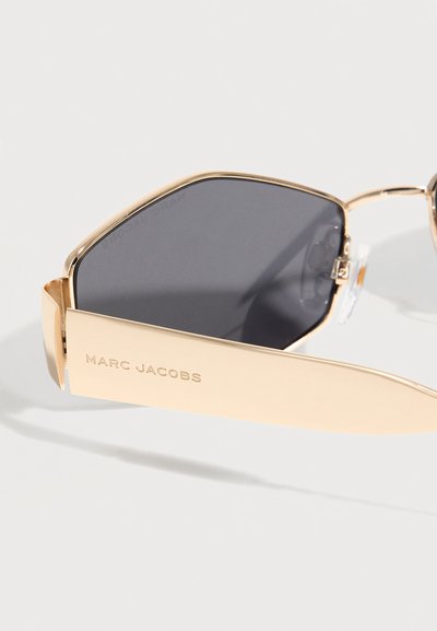 Γυαλιά ηλίου με χρυσή κορνίζα και σκούρα φακούς, καθώς και φαρδιές χρυσές πλευρές με χαραγμένο το "MARC JACOBS" σε ανοιχτό φόντο.