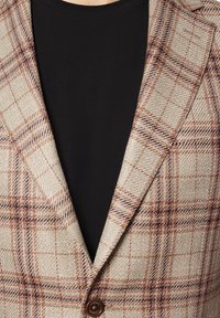 Beżowa marynarka w kratę z brązowymi i granatowymi akcentami, teksturowana tkanina, wyłogi w kształcie szpicu oraz zapięcie na jeden guziczek na czarnej koszuli.