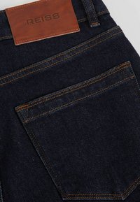 Tasca posteriore di jeans in denim blu scuro con toppa in pelle marrone etichettata "REISS" e dettagli di cuciture arancioni.