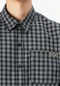 Black and white plaid button-up shirt with a collared neck, front pocket, and logo tag. Fabric appears soft with a textured pattern.