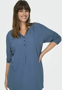 Túnica azul de manga larga con un ajuste relajado, que presenta un escote abotonado, dos bolsillos frontales y un tejido suave y texturizado.