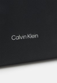Superficie in pelle nera con texture che presenta il logo "Calvin Klein" in argento, posizionato in modo prominente. Design minimalista senza ulteriori decorazioni.