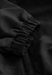 Tissu noir à texture lisse, avec un poignet élastique froncé. Le matériau semble léger et résistant à l'eau, avec une fermeture éclair dissimulée.